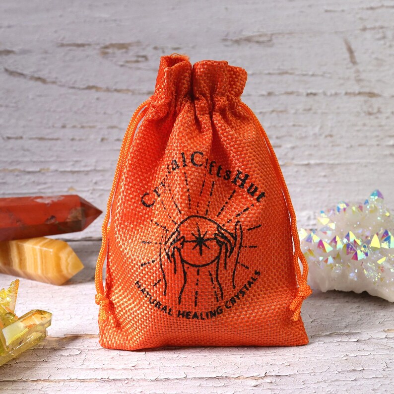 May include: An orange drawstring bag with the text "Crystal Gifts Hut" and "Natural Healing Crystals" printed in black. The bag is made of a textured material and is filled with crystals. Other crystals are visible in the background.