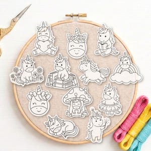 Puede incluir: Un bastidor de bordado redondo de madera que contiene un panel de tela beige con doce pegatinas de unicornios en blanco y negro. Las pegatinas muestran varios unicornios sentados, leyendo, con tutú o descansando bajo una seta. Tijeras visibles.