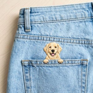 Peut inclure: Un vêtement en tissu denim bleu clair doté d'une poche arrière avec un chiot golden retriever brodé. Le chien a un pelage beige clair, une truffe noire et une gueule ouverte montrant une langue rose, ses pattes avant posées sur le bord.