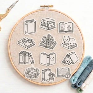 Könnte beinhalten: Eine Sammlung von schwarz-weißen Aufklebern zum Thema Bücher, angeordnet auf einem beigen Stoff in einem Stickrahmen. Die Aufkleber zeigen verschiedene Buchdesigns, darunter Stapel, offene Bücher mit Herzen, Blumen und eine Katze. Stickgarn in Blau- und Weißtönen ist sichtbar.