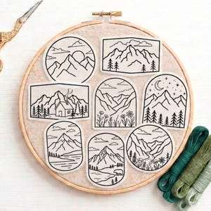 May include: A wooden embroidery hoop containing eight white fabric patches with black line art illustrations of mountain landscapes, forests, cabins, rivers, and night skies, accompanied by three skeins of green embroidery thread.