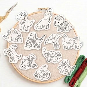 May include: A collection of twelve black and white dinosaur stickers featuring holiday themes, arranged on a round wooden embroidery hoop with beige fabric. The designs include dinosaurs with scarves, hats, Christmas trees, gifts, and a gingerbread man.