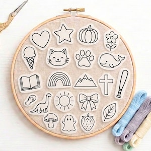 Peut inclure: Un tambour à broder rond en bois contenant un panneau en tissu beige avec vingt écussons individuels en noir et blanc. Les motifs incluent un cœur, une étoile, une citrouille, une fleur, une glace, un chat, une patte, une baleine, un livre, un arc-en-ciel, une montagne, une croix, une batte de baseball, une tong, un dinosaure, un soleil, un nœud, un champignon, un fantôme, une fraise et une feuille.