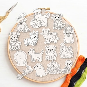 May include: A round wooden embroidery hoop containing a beige fabric panel with fifteen black and white line art stickers of dogs in Halloween costumes, including a mummy dog, a skeleton dog, a ghost dog, and dogs with pumpkins and witch hats.