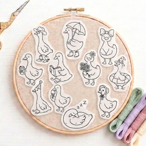 May include: A selection of quirky duck stickers, each depicted in different scenarios, such as reading a book or wearing a party hat. The stickers are displayed on a beige embroidery hoop, with embroidery threads in shades of pink, purple, and green.
