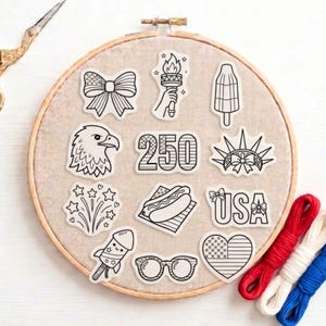 May include: A collection of black and white stickers with patriotic themes, including a bow, torch, ice pop, eagle, fireworks, hot dog, and the word "USA". The stickers are arranged on a beige fabric within an embroidery hoop, with red, white, and blue embroidery floss.