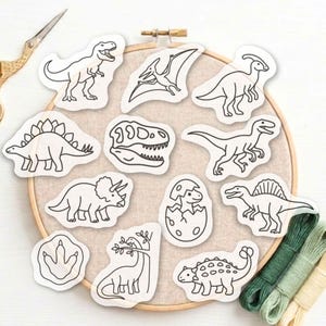 May include: A wooden embroidery hoop containing light-colored fabric with eleven black and white dinosaur-themed iron-on patches. The designs include various dinosaurs, a skull, a footprint, and a hatchling. Gold scissors and three green embroidery threads are visible.