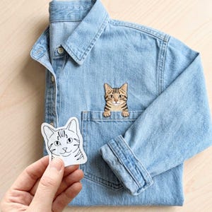 May include: A light blue denim button-down shirt featuring an embroidered brown tabby cat peeking out from the chest pocket. A separate white sticker with a black line drawing of a cat face is held in the foreground, showing a matching feline design.