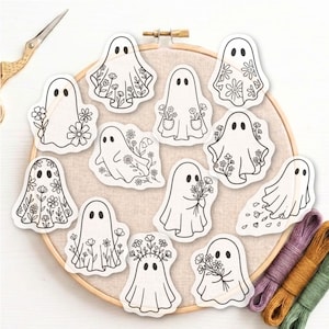 Può includere: Una collezione di dodici adesivi a forma di fantasma bianco con contorni neri, ognuno caratterizzato da motivi floreali, bouquet o tasche unici. Gli adesivi sono disposti su un telaio da ricamo circolare in legno con tessuto beige.