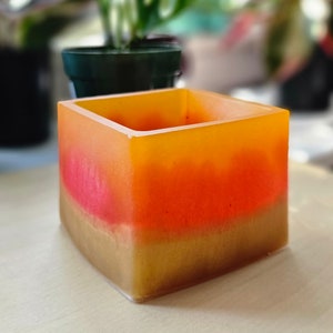 May include: A square-shaped planter with a layered design. The bottom layer is a light brown color, the middle layer is a bright orange color, and the top layer is a light yellow color.