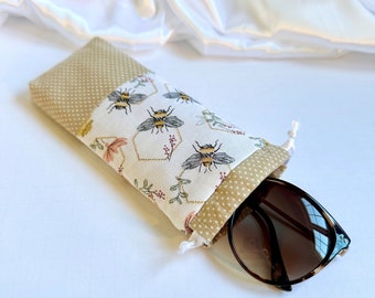 May include: A beige and white fabric eyeglass case with a drawstring closure. The case features a pattern of bees and honeycombs. A pair of brown sunglasses is inside the case.