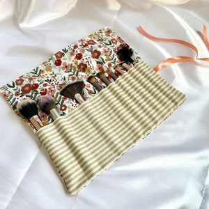 May include: A floral patterned makeup brush roll with a striped fabric lining. The roll is rolled up and has several makeup brushes inside.