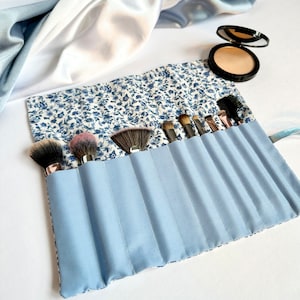 May include: A blue and white floral makeup brush roll with eight slots for brushes. The roll is rolled up and has a blue fabric exterior with white floral print. The roll is on a white surface.