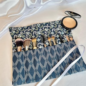 May include: A navy blue and white floral print makeup brush roll with a diamond pattern on the front. The roll contains various makeup brushes and a compact powder. A white ribbon is tied around the roll.
