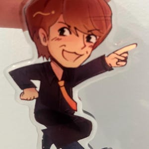 Puede incluir: Un llavero acrílico transparente con una ilustración de dibujos animados de un hombre con cabello castaño, que lleva un traje negro, una corbata naranja y pantalones azules. El hombre está señalando con una sonrisa. El llavero está sujeto por una cuerda negra.