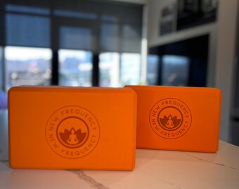 May include: Two orange yoga blocks with the text "NEW FREQUENCY" and a lotus flower logo. The blocks are rectangular and sit on a white countertop. The background includes a window and a glimpse of a cityscape.