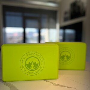 May include: Two lime green yoga blocks with the text "NEW FREQUENCY" and a lotus flower logo. The blocks are rectangular and appear to be made of foam. The blocks are on a white marble surface.