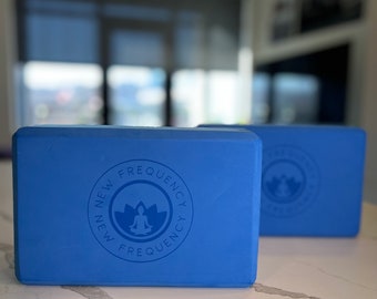 May include: Two blue yoga blocks with rounded edges are displayed on a white countertop. Each block has a circular logo with the text "NEW FREQUENCY" and a meditating figure. The blocks are designed for yoga and fitness.