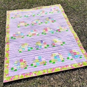 May include: A handmade quilt with a lavender background and a repeating pattern of colorful patchwork turtles. The quilt has a green and pink floral border and is lying on grass.