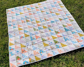 May include: A patchwork quilt with a white background and a pattern of colourful triangles. The quilt is made of various fabrics with different patterns, including floral, geometric, and abstract designs. The quilt is laid out on a green grassy surface.