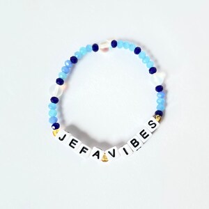 May include: A blue and white beaded bracelet with the word "Jefavibes" spelled out in white letters on white beads.