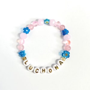 May include: A pink and blue beaded bracelet with flower charms and the name "LUCHONA" spelled out in white letters with gold accents.