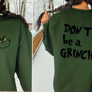 May include: Dark green sweatshirt featuring a Grinch face on the front and the text "DON'T be a GRINCH" on the back. The Grinch's face is black with yellow eyes. The sweatshirt is a solid color and has a crew neck.