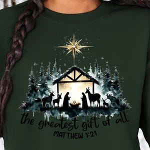 May include: Dark green sweatshirt featuring a Nativity scene with a star, trees, and the text "the greatest gift of all" and "Matthew 1:21". The design is in a watercolor style.