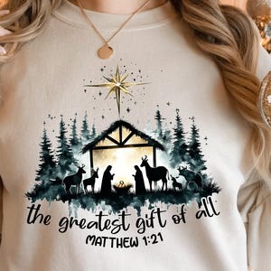 May include: Cream-colored sweatshirt with a Nativity scene graphic. The design features a silhouette of the Holy Family, animals, and a star above a stable, with the text "the greatest gift of all" and Matthew 1:21.