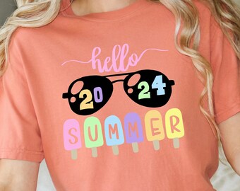 May include: A coral colored t-shirt with the text "hello summer" in pink and black. The text is surrounded by a pair of sunglasses with the number "2024" on the lenses.  There are six colorful popsicle graphics below the sunglasses.