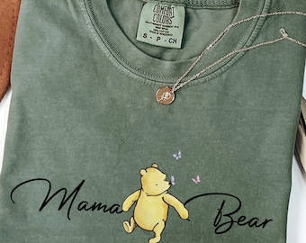 May include: A green t-shirt with a yellow cartoon bear and the text "Mama Bear" in black. The bear is blowing on a dandelion with purple and pink flowers.