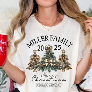May include: A cream-coloured t-shirt featuring a Christmas tree design and the text "MILLER FAMILY 2025 Merry Christmas Colorado Springs CO." The design includes three Christmas trees with leopard print bows. A red mug with "oh what FUN!" is held.