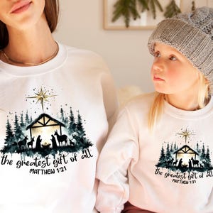 May include: White sweatshirts with a Christmas-themed graphic. The design features a nativity scene with a star, trees, and the text "the greatest gift of all" and "Matthew 1:21". One sweatshirt is worn by a child.