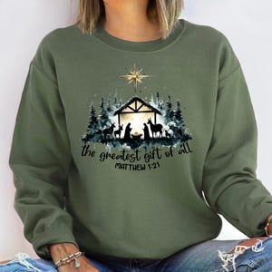 May include: Olive green sweatshirt with a graphic of a nativity scene, including a star, stable, and silhouettes of animals and figures. The text "the greatest gift of all" and "MATTHEW 1:21" are printed below the graphic.