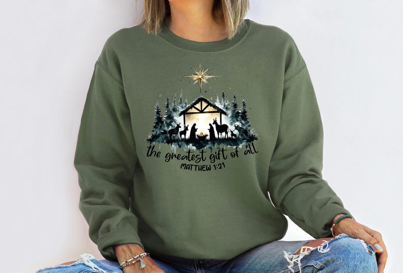 May include: Olive green sweatshirt with a graphic of a nativity scene, including a star, stable, and silhouettes of animals and figures. The text "the greatest gift of all" and "MATTHEW 1:21" are printed below the graphic.