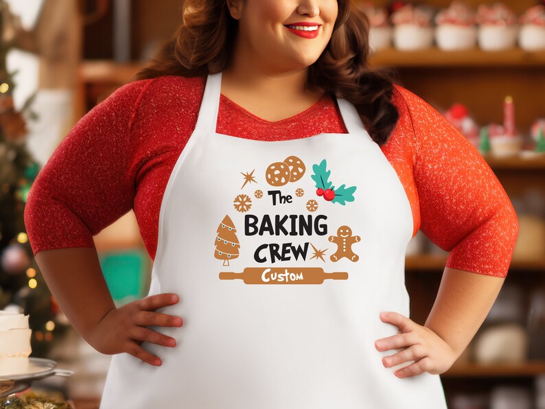 Puede incluir: Delantal blanco con un hombre de jengibre marr&oacute;n, un &aacute;rbol de Navidad y un rodillo. El texto en el delantal dice "The Baking Crew Custom".