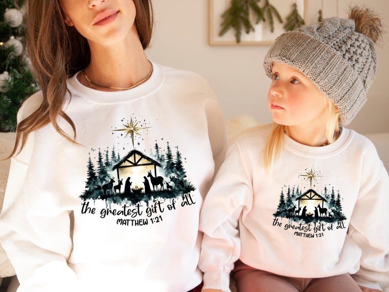May include: White sweatshirts with a Christmas-themed graphic. The design features a nativity scene with a star, trees, and the text "the greatest gift of all" and "Matthew 1:21". One sweatshirt is worn by a child.