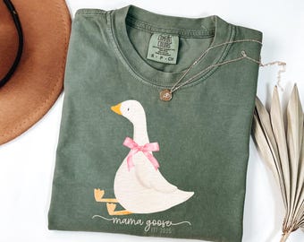 May include: A green t-shirt with a white goose wearing a pink bow. The goose is sitting on a branch and the text "mama goose est 2025" is printed below it.