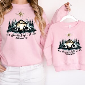 May include: Pink sweatshirts with a Christmas Nativity scene graphic. The design includes a star, a stable, silhouettes of animals and people, and the text "the greatest gift of all" and "Matthew 1:21".