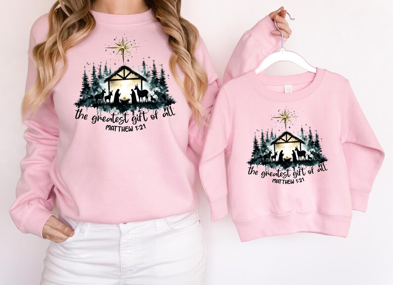 May include: Pink sweatshirts with a Christmas Nativity scene graphic. The design includes a star, a stable, silhouettes of animals and people, and the text "the greatest gift of all" and "Matthew 1:21".