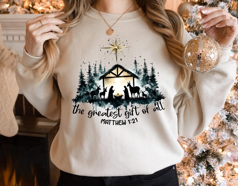 May include: Cream-colored sweatshirt with a Nativity scene graphic. The design features a silhouette of the Holy Family, animals, and a star above a stable, with the text "the greatest gift of all" and Matthew 1:21.