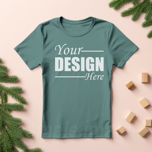 May include: A teal-colored t-shirt with the text "Your DESIGN Here" in white. The shirt is displayed on a light pink surface, with wooden blocks and evergreen branches in the background.