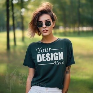 May include: A woman wearing sunglasses and a dark green t-shirt with the text "Your Design Here" printed on the front. She is standing in a park with green trees in the background.