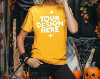 May include: A woman wearing a yellow t-shirt with the text "YOUR DESIGN HERE" in white. The shirt has a crew neck and short sleeves. The woman is standing in front of a dark background with autumn foliage and pumpkins.
