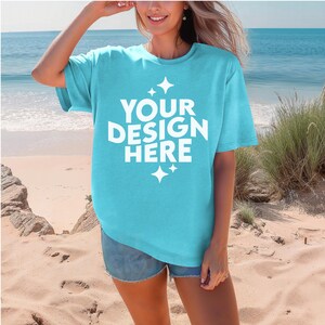 Puede incluir: Una camiseta azul claro de manga corta con un gráfico blanco que dice "YOUR DESIGN HERE" con estrellas a cada lado del texto. La modelo lleva la camiseta con pantalones cortos de mezclilla y está de pie en una playa de arena.