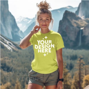 Può includere: Una donna indossa una maglietta gialla brillante con il testo "YOUR DESIGN HERE" in bianco. La maglietta ha un collo rotondo e maniche corte. La donna è in piedi davanti a un paesaggio montano.