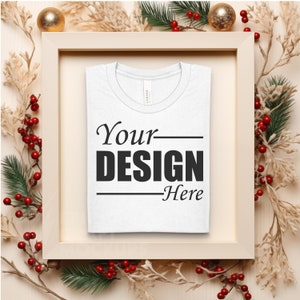Può includere: Maglietta bianca con la scritta "Your Design Here" stampata sul davanti. La maglietta è esposta in una cornice di legno con decorazioni natalizie.