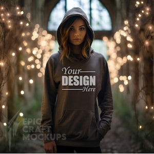May include: A gray hooded sweatshirt with the text "Your DESIGN Here" printed on the front. The model is wearing the sweatshirt with the hood up and is standing in a dimly lit room with string lights.