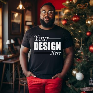 Puede incluir: Una camiseta negra con el texto "Your DESIGN Here" en letras blancas. La persona que lleva la camiseta está de pie frente a un árbol de Navidad.