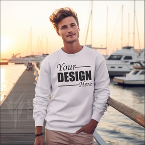 May include: White crew neck sweatshirt with the text "Your DESIGN Here" in black. The sweatshirt is a solid white color and has long sleeves. The background shows a dock with boats and a sunset.